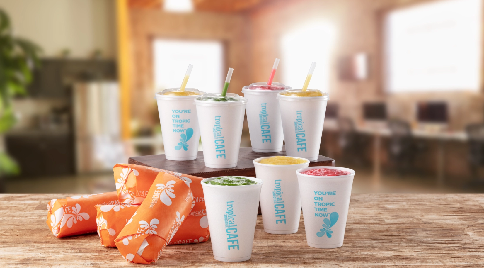 4 Tropical Smoothie Cafe wraps and 7 smoothies stacked on a table with a blurry background of a room.