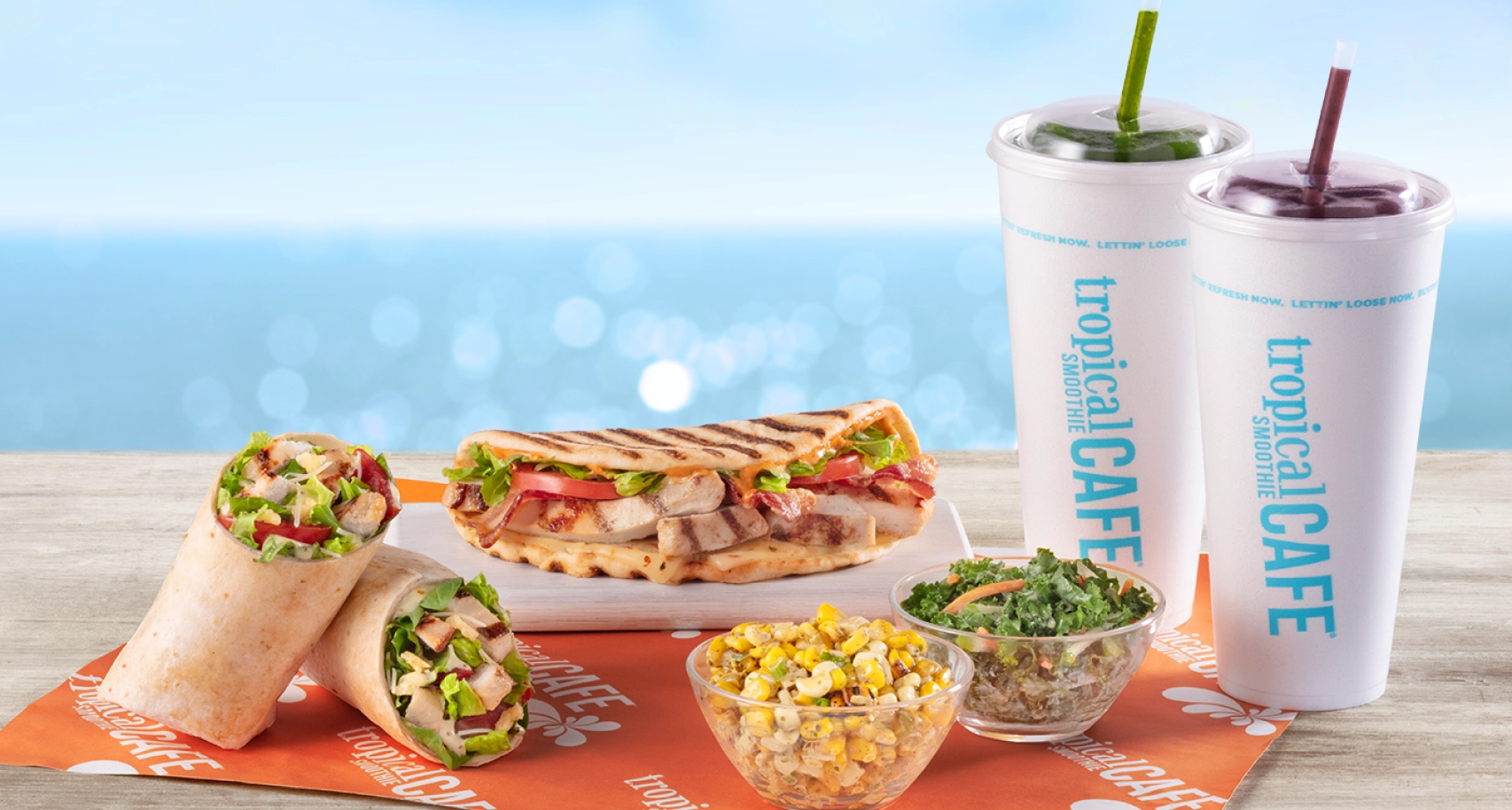 Delicious Tropical Smoothie Cafe menu items - 2 large smoothies, Tropical Smoothie Cafe flatbread, grilled chicken wrap, jalapeno corn and kale n apple slaw placed on a serving paper on the beach.