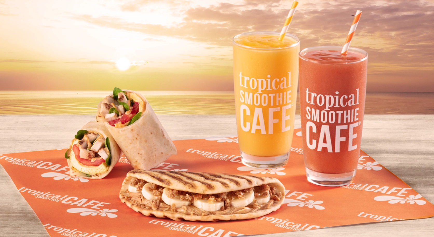 Tropical Smoothie Cafe Smoothies, Flatbread and wrap placed on an orange serving paper with sun setting in the background.